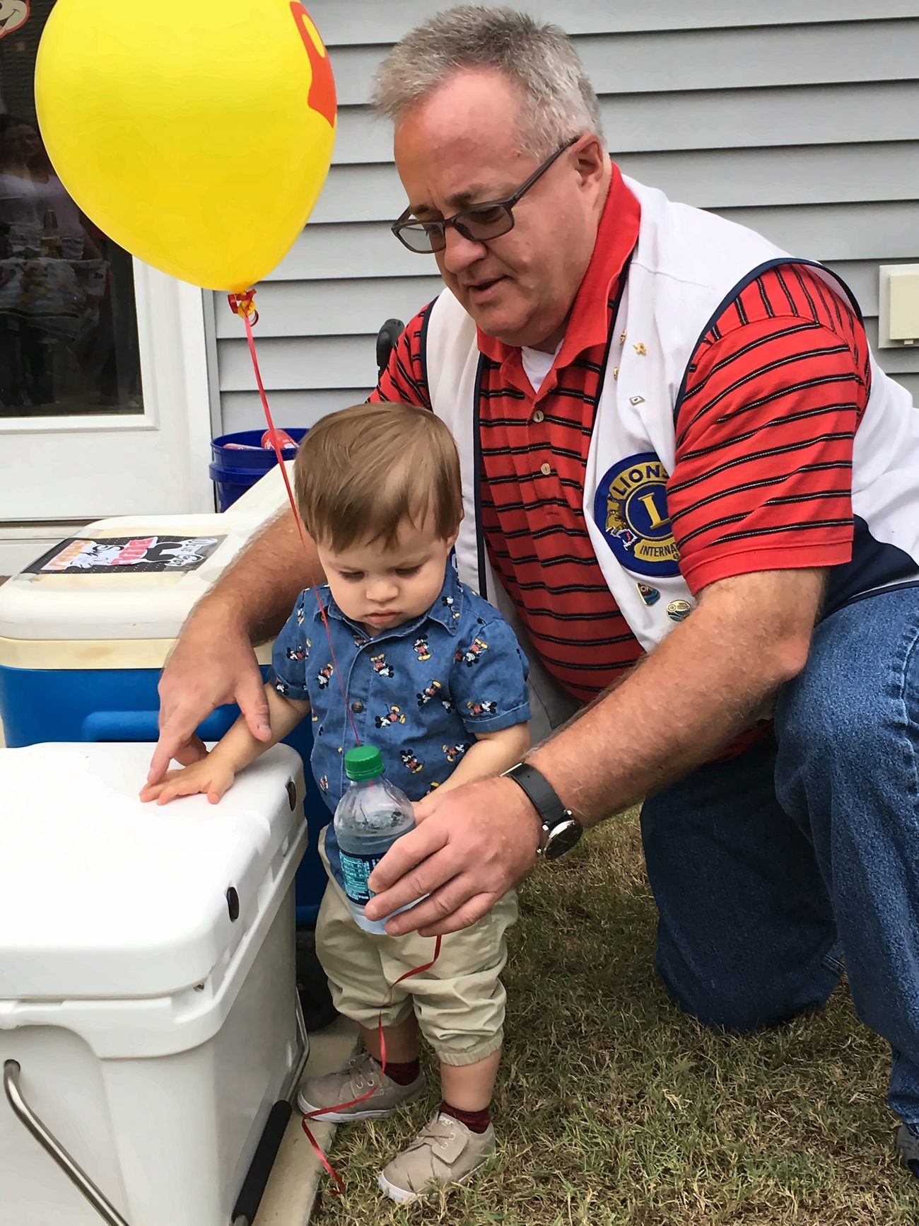 A Lexington Lions Club member kneeling beside a toddler holding a yellow balloon at a community event.