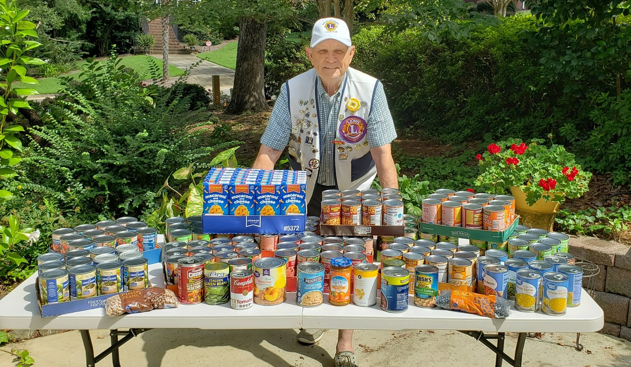 A Lexington Lions Club member in club cap and vest standing behind a table piled high with canned goods collected for a district food drive.