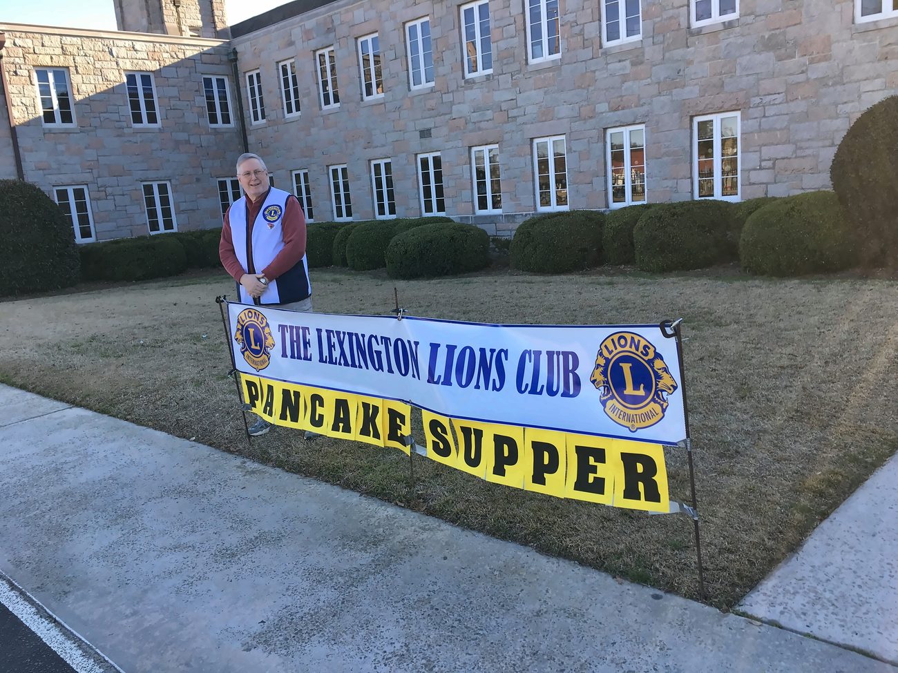 A Lexington Lions Club member standing beside the club's Pancake Supper banner outside a community building.
