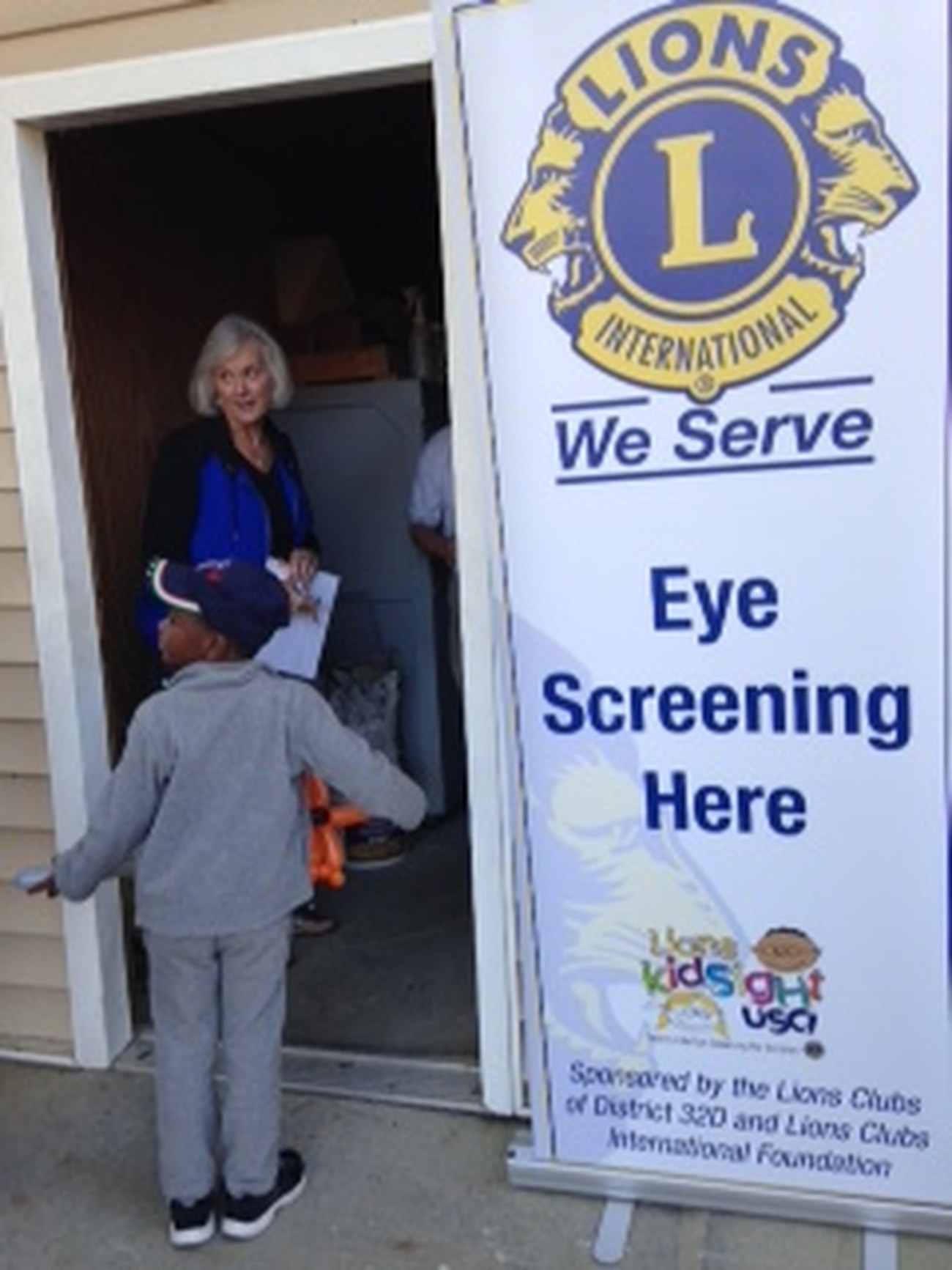 A Lexington Lions Club volunteer greets a child beneath a 'We Serve, Eye Screening Here' banner at a community vision-screening event.