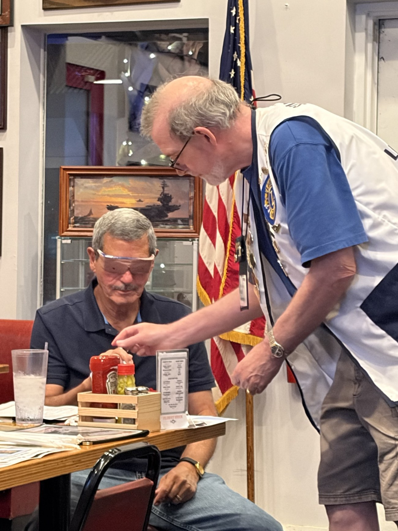 Lion John Reilly guiding a seated Lexington Lions Club member through a reading exercise while the member wears vision-impairment simulation glasses, during a Weeks of Service: Vision presentation.